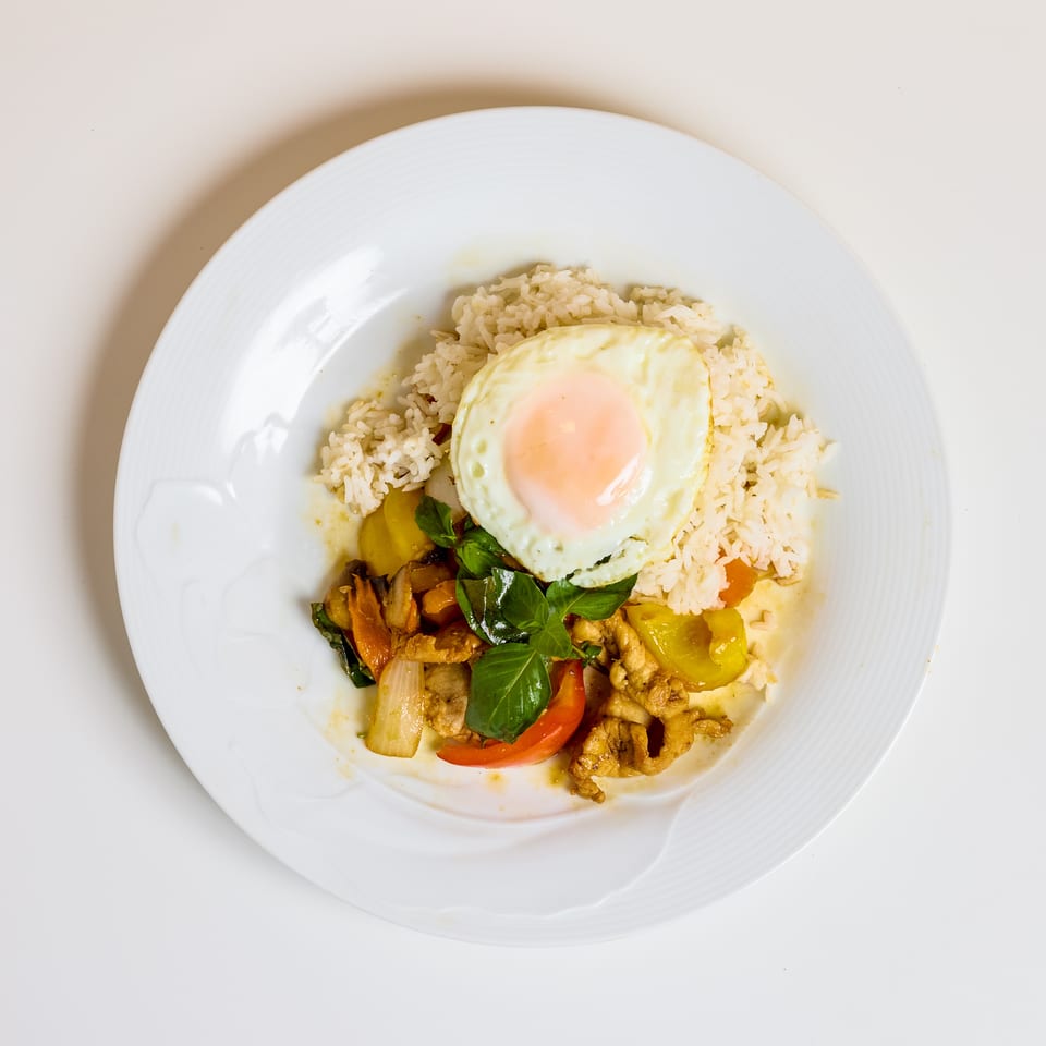 Thai-style stir-fried chicken with vegetables, basil, and jasmine rice topped with a sunny-side-up egg on a white plate.