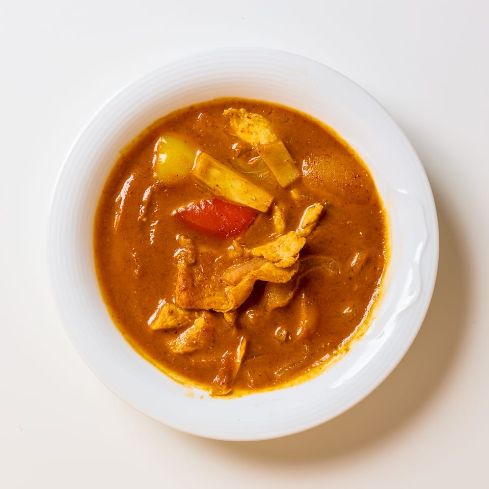 Bowl of rich brown chicken curry with vegetables, including potatoes, peppers, and leeks, served on a white plate.