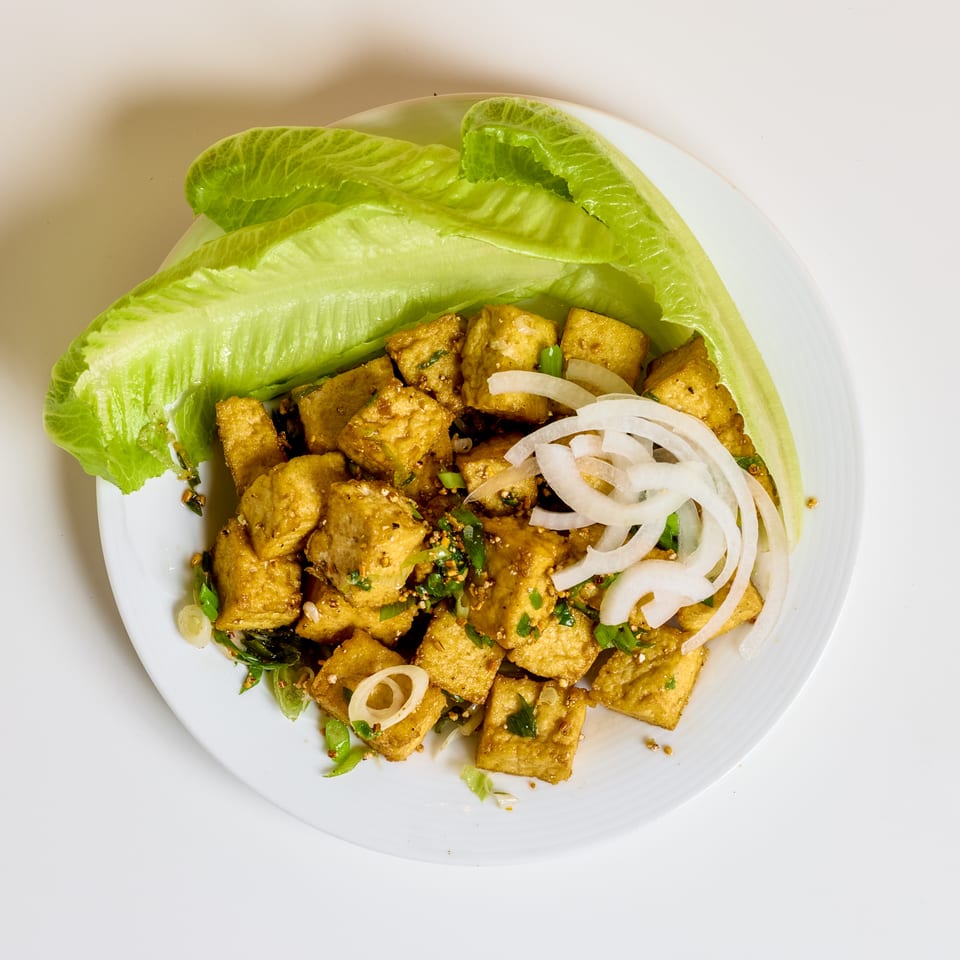 Seasoned golden tofu cubes garnished with sliced onions, herbs, and lettuce leaves on a white plate.