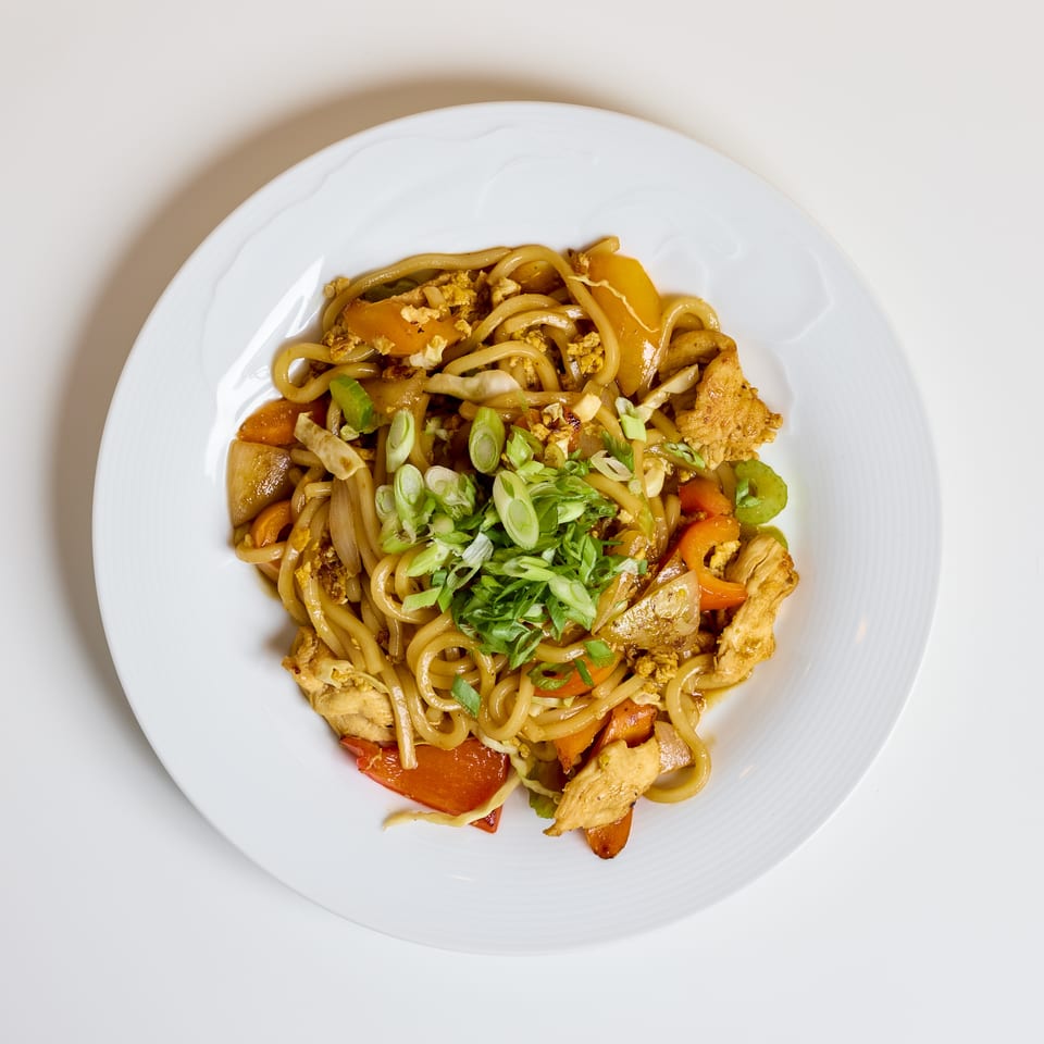 Stir-fried noodles with chicken, vegetables, scrambled egg, and sliced green onions on a white plate.