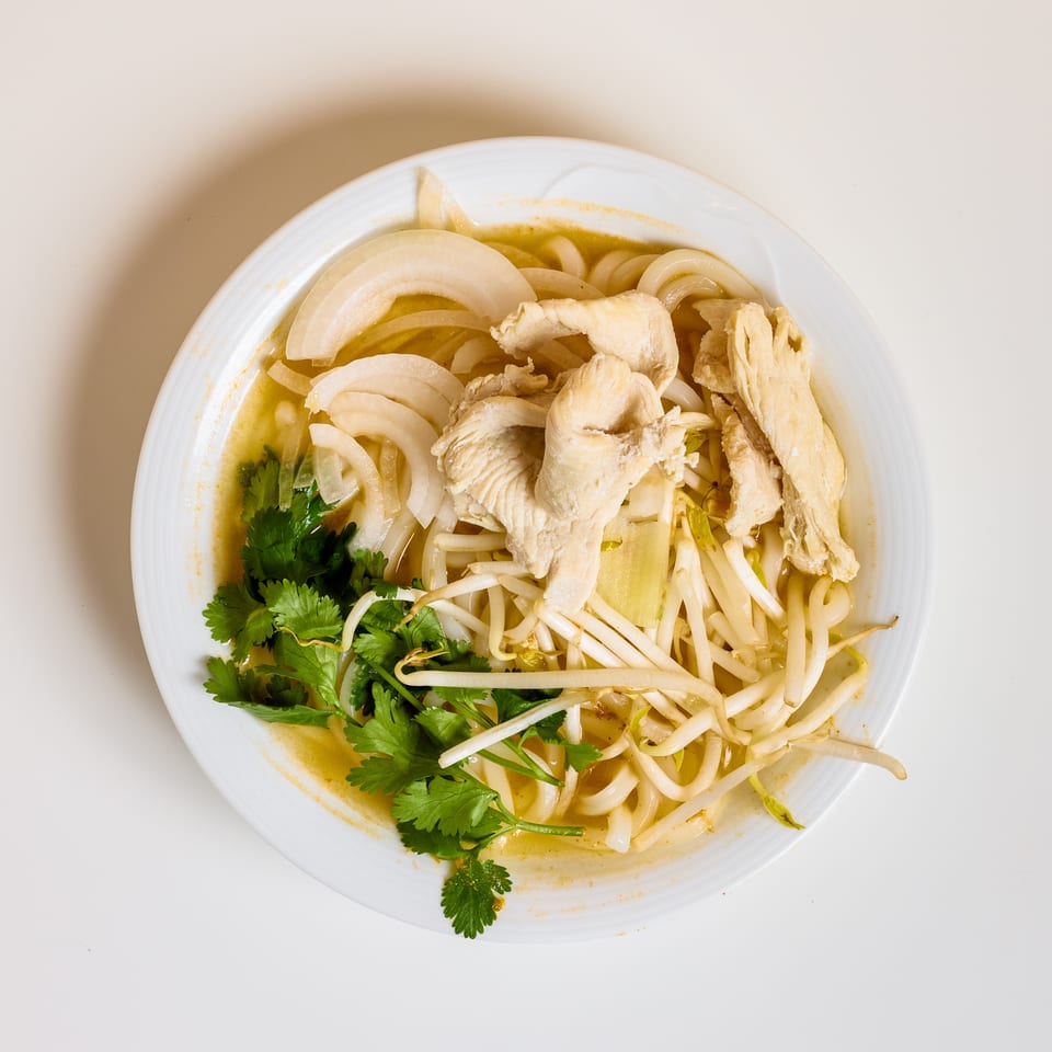 Bowl of noodles in broth topped with sliced chicken, onions, bean sprouts, cilantro, and vegetables.