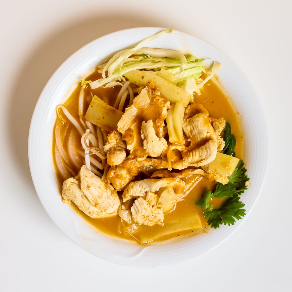 Bowl of chicken in golden curry broth with bamboo shoots, bean sprouts, cabbage, and fresh herbs.