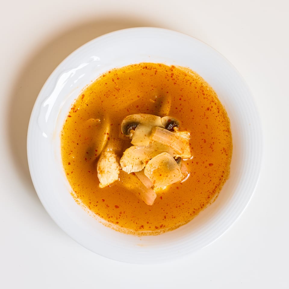 Bowl of orange curry soup with sliced chicken, mushrooms, and onions on a white plate.
