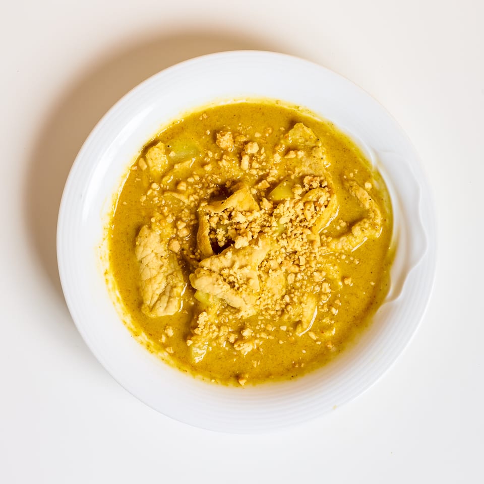 Overhead view of creamy golden curry with chunks of meat and vegetables, topped with crushed nuts, in a white bowl.