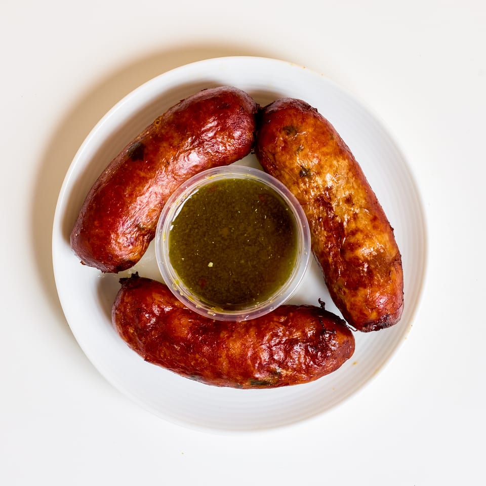 Three grilled sausages surround a cup of green dipping sauce on a white plate.