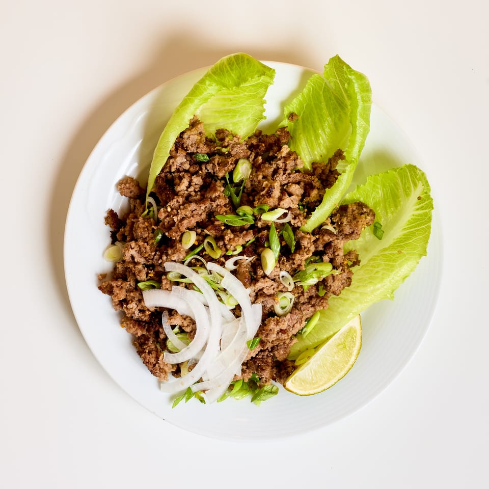 Seasoned minced meat lettuce wraps garnished with sliced onions, scallions, herbs, and lime wedges on a white plate.