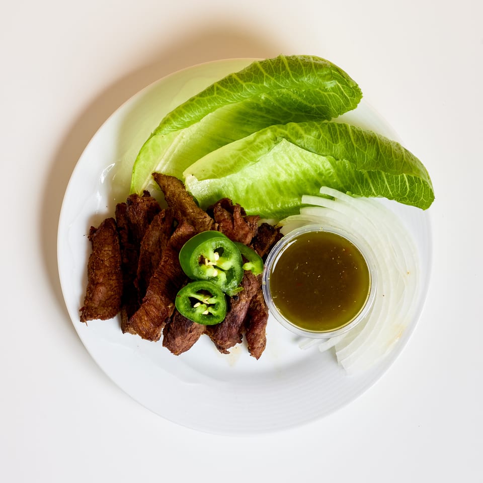 Plate of grilled meat strips with romaine lettuce, sliced onion, jalapeños, and dipping sauce.