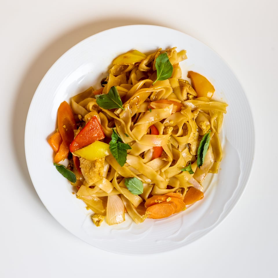 Plate of stir-fried flat noodles with chicken, colorful vegetables, and fresh basil leaves.