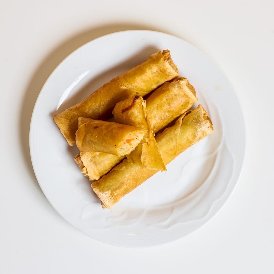 Golden fried spring rolls and samosas arranged on a white plate, with one spring roll cut open to reveal vegetable filling.